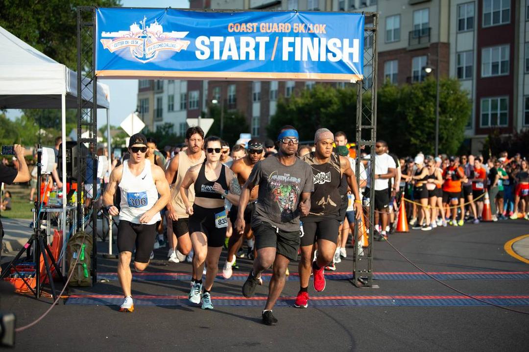Runners bursting across the mat under the Coast Guard 5K/10K start and finish banner in Portsmouth